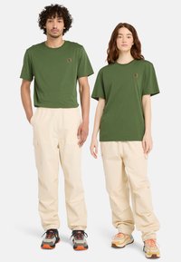 Gröna bomullströjor, löst sittande khaki cargobyxor och flerfärgade sneakers på två modeller. Har en logotyp-patch på tröjorna.
