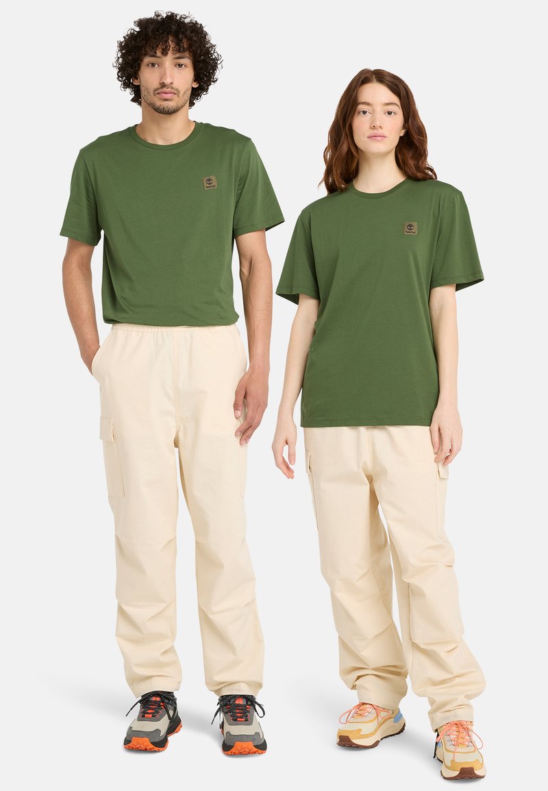 Gröna bomullströjor, löst sittande khaki cargobyxor och flerfärgade sneakers på två modeller. Har en logotyp-patch på tröjorna.