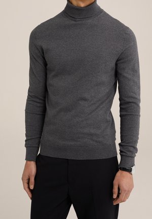 Mann trägt einen taillierten dunkelgrauen Rollkragenpullover mit langen Ärmeln und schwarze Hosen, und zeigt eine silberne Armbanduhr am linken Handgelenk.