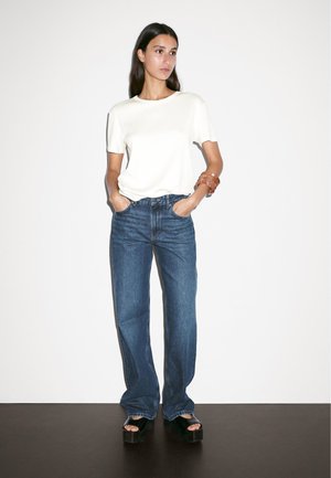 Maglietta bianca a maniche corte abbinata a jeans blu a vita alta con gamba dritta e sandali platform neri, su sfondo neutro.