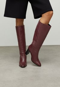 Bottes montantes en cuir bordeaux avec un bout pointu et un talon bloc, présentant une texture lisse et un design simple.