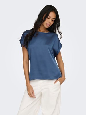 Femme aux longs cheveux foncés portant un chemisier bleu ample à manches courtes et un pantalon blanc à jambes larges, debout avec une main dans la poche.