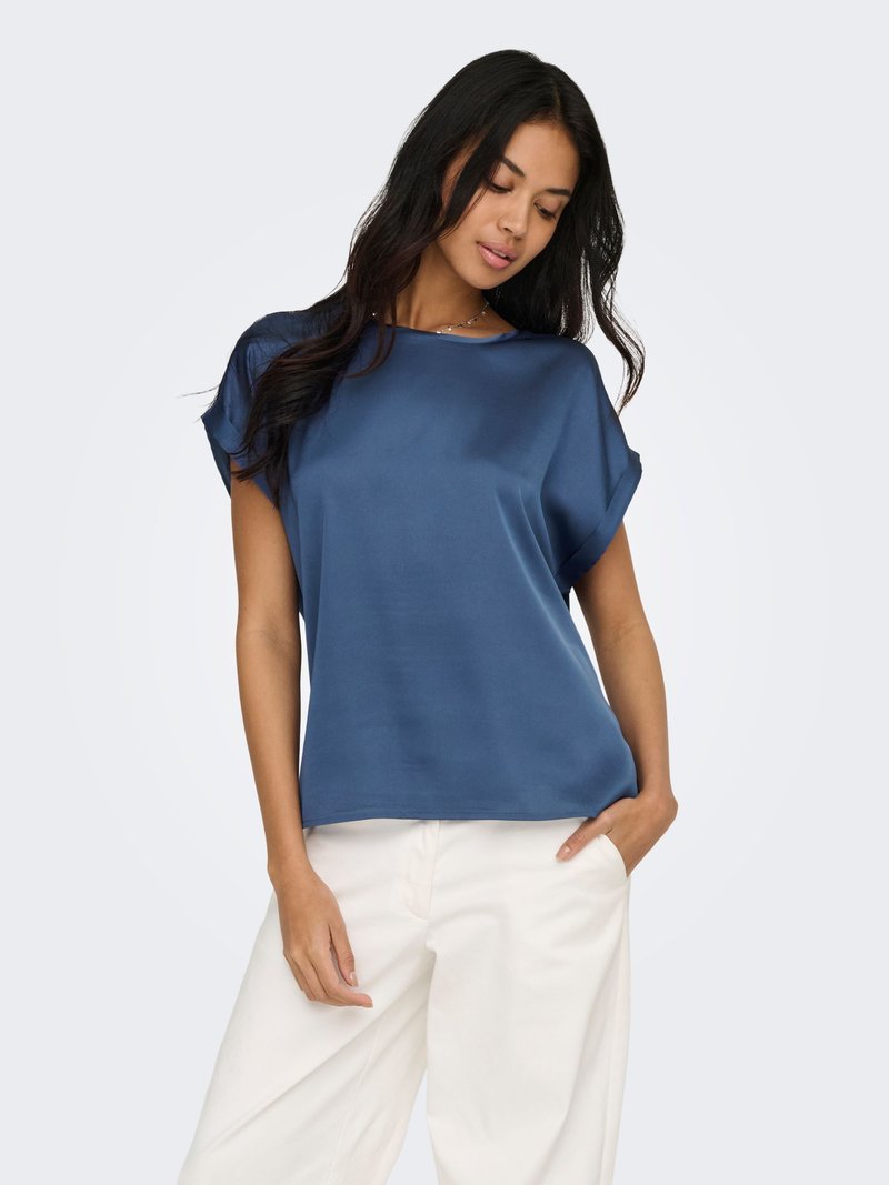 Femme aux longs cheveux foncés portant un chemisier bleu ample à manches courtes et un pantalon blanc à jambes larges, debout avec une main dans la poche.