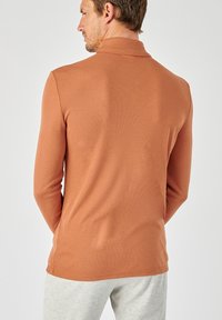 Langwerpige, roestkleurige pullover met een ribbeltextuur en een mock-kraag, gedragen over lichtgrijze, gestructureerde broek met een fitted design.