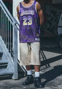 Fioletowa koszulka koszykarska z napisem "Lakers" i numerem "23", w zestawieniu z khaki szortami i czarnymi wysokimi trampkami. Scena z schodami i graffiti.