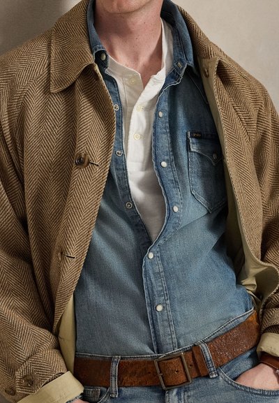 Man draagt gelaagde kleding waaronder een bruine visgraat jas, denim overhemd, wit henley shirt en bruine leren riem.
