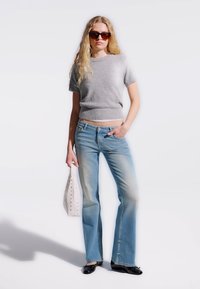 Mlada ženska z rjavimi sončnimi očali, sivim puloverjem s kratkimi rokavi, zbledelimi modrimi kavbojkami, črnimi čevlji in belim ročnim kovčkom.