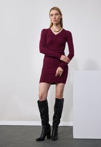Robe tricot à manches longues bordeaux avec texture côtelée et décolleté en V, associée à des bottes noires montantes à talon épais.