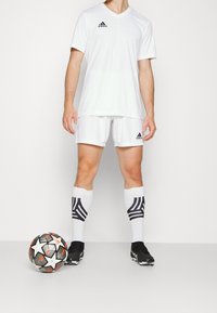 Camiseta y pantalones cortos de fútbol Adidas blancos con detalles en negro, combinados con calcetas blancas hasta la rodilla que presentan rayas negras, y un balón de fútbol con un diseño llamativo.