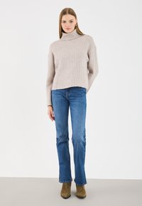 Pulover bej deschis cu guler înalt și nervuri, asortat cu blugi evazați albaștri și botine maro până la glezne. Textura pare moale; puloverul are un design scurt.