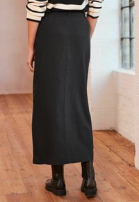 Falda maxi negra confeccionada en una tela suave, con un corte recto. El diseño incluye una cinturilla y bolsillos traseros, combinada con botas negras.