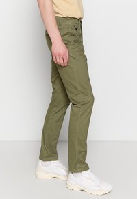 Hombre vestido con pantalones verde oliva y zapatillas blancas, posando frente a un fondo gris claro liso.