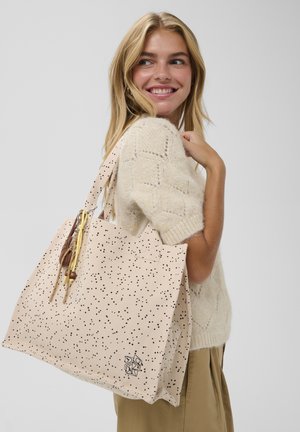 Bolsa tote de lona beige con motas negras, que cuenta con dos asas y borlas decorativas. Incluye un pequeño logo en el frente.
