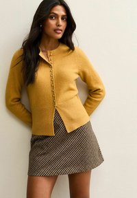 Mosterdkleurige cardigan met voor knopen, ribbelzoom; gecombineerd met een zwart en bruin geruit minirok. Gladde textuur, aangesloten ontwerp.