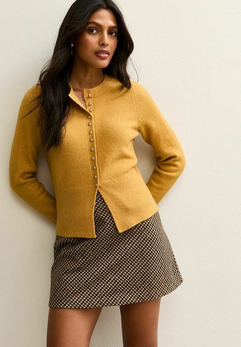 Mosterdkleurige cardigan met voor knopen, ribbelzoom; gecombineerd met een zwart en bruin geruit minirok. Gladde textuur, aangesloten ontwerp.