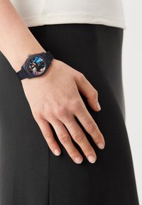 Black silicone watch with a textured strap, round face featuring colorful graphics and numerals, worn on a wrist against a black fabric background.