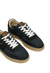 Tmavě modré tenisky z měkkého materiálu a broušené kůže, s bílými tkaničkami, hnědým koženým detailem na jazyku a gumovou podrážkou.