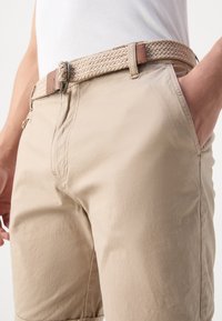 Pantaloni corti beige con cintura intrecciata, dotata di una fibbia in metallo e un accento in pelle. Il tessuto è liscio con cuciture visibili e tasche.