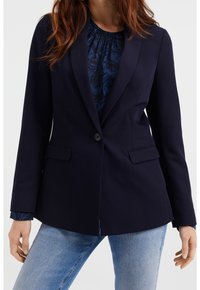 Blazer bleu marine en tissu lisse avec une fermeture à un bouton, des revers crantés et deux poches avant, porté sur un chemisier à motifs.