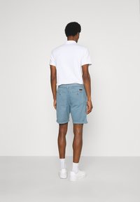 Pantalones cortos de algodón azul claro con dobladillos enrollados, bolsillos traseros y un sutil detalle de etiqueta de la marca. Camiseta tipo polo blanca de manga corta y zapatillas.