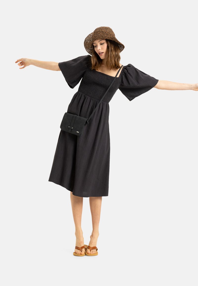 Vestido negro con mangas abullonadas y un cuerpo fruncido, combinado con un bolso negro bandolera y sandalias de tira marrón. La modelo posa con los brazos extendidos.