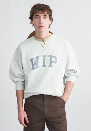 Felpa grigio chiaro con la scritta "WIP" stampata in grigio scuro. Presenta uno scollo rotondo e polsini larghi. Abbinata a pantaloni marroni.