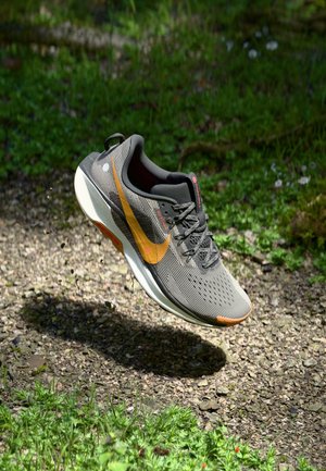 Gray and orange Nike running shoe mid-air over gravel path with green grass background, showing side and bottom sole details.