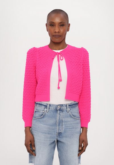 Cardigan scurt de culoare roz aprins, cu un model texturat, mâneci bufante și detaliu cu fundă la decolteu, asortat cu blugi din denim de culoare albastru deschis.