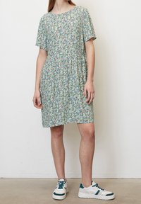 Mladá osoba oblečená v krátkém rukávovém šatech s pastelovým květinovým vzorem a bílých teniskách s tmavě zelenými akcenty, stojící před bílou zdí.