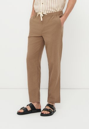 Άτομο που φορά καφέ παντελόνι με κορδόνι, ριγέ πουκάμισο μέσα στην παντελόνα και μαύρα σανδάλια με διπλό λουράκι, στέκεται μπροστά σε λευκό φόντο.