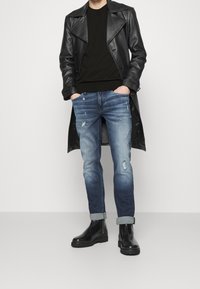 Trench-coat en cuir noir, pull noir, jean bleu délavé avec des détails usés, revers retroussés, et bottines noires brillantes.