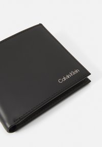 Juodas odinis piniginė su siuvinėtais kraštais ir įspaustu sidabriniu "Calvin Klein" logotipu. Siūlo minimalistinį, sulankstomą dizainą.