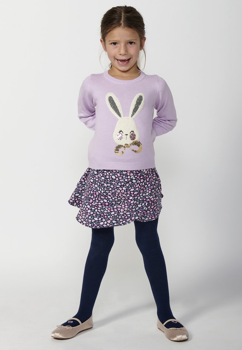 Lila neule, jossa on pupun kuvio, kukkamekko, tummansiniset sukkahousut ja beige ballerina-tossut. Huomattavia kimaltavia yksityiskohtia pupussa.