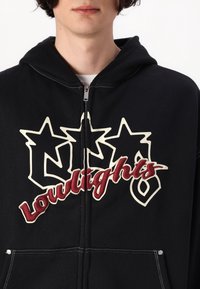 Sweat à capuche noir zippé avec de grands motifs graphiques blancs et rouges, comprenant des étoiles et le mot "Loudights", avec des coutures contrastantes et des poches.