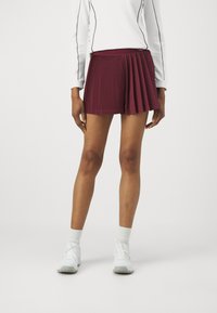 Mörkröd plisserad kjol med ett åtsittande midjeband, tillsammans med vita sportskor och strumpor. Materialet verkar slätt med tydliga plisseringar.