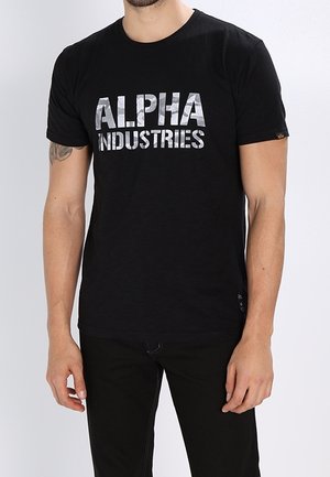 Mężczyzna w czarnej koszulce z srebrnym napisem „ALPHA INDUSTRIES” oraz w czarnych spodniach, stojący z ramionami luźno opuszczonymi wzdłuż ciała.