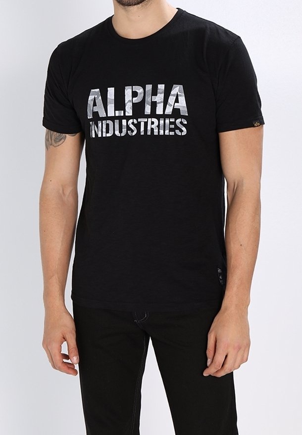 Homme portant un t-shirt noir avec un texte argenté "ALPHA INDUSTRIES" et un pantalon noir, debout les bras détendus le long du corps.