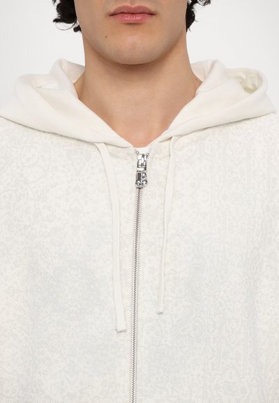 Hoodie blanc zippé avec un motif texturé, doté d'une capuche à cordon de serrage et d'une fermeture éclair en argent. Tissu doux avec un détail de design subtil.