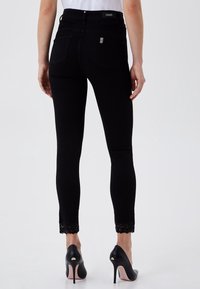 Jeans skinny negros con cintura alta, con detalle de encaje en el tobillo, bolsillos traseros y una etiqueta de logo en la cinturilla. Combinados con tacones negros.