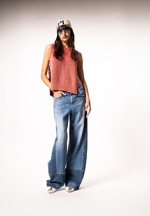 Žena oblečená v červenom pletenom top bez rukávov, široké bledomodré džíny s vyblednutím, biele sandále s otvorenou špičkou, béžový šál na hlave s čiernym vzorom a modré slnečné okuliare.