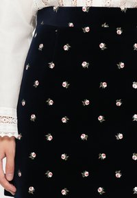 Biała bluzka z koronkowymi rękawami, włożona w czarną spódnicę z małym różowym i białym haftem kwiatowym, z lewą ręką opartą na boku.