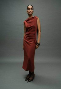 Rostrote Midikleid mit drapiertem Ausschnitt und asymmetrischem Design. Glatter Stoff, figurbetonender Schnitt, ergänzt durch hohe dunkle Absätze und Armreifen.