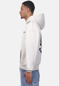 Light beige oversized hoodie with a large graphic on the sleeve, featuring a drawstring hood and front pocket. Soft material and ribbed cuffs.