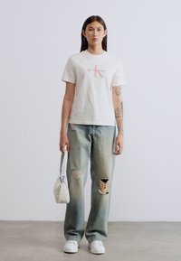 Άσπρο κοντομάνικο T-shirt με κόκκινο λογότυπο, συνδυασμένο με ανοιχτό μπλε φαρδιά τζιν με φθαρμένες περιοχές. Αξεσουάρ περιλαμβάνουν μια μικρή λευκή τσάντα.