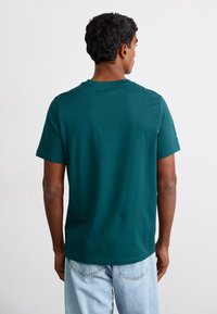 Mørk teal kortærmet t-shirt i bomuldsstof, med rund hals og en afslappet pasform. Modellen har lyseblå denimjeans på.