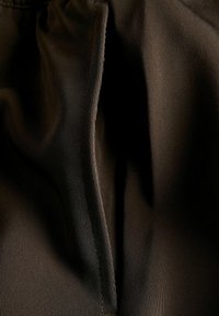 Tumši brūnas auduma daļa ar gludu tekstūru un plecu; tīrā šuvuma līnija gar malu izceļ auduma struktūru un kritumu.