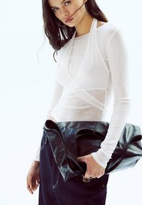 White long-sleeve top features a wrap design. The model holds a black leather bag with a smooth texture and oversized shape. Dark denim jeans are worn.