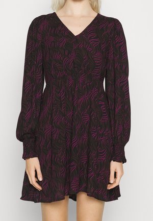 Femme portant une robe noire à manches longues avec des lignes ondulées abstraites roses, encolure en V, taille élastique et poignets froncés.