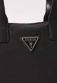 Borsa nera realizzata in nylon con una texture liscia. Presenta una targhetta con logo triangolare in oro e nero con la scritta "GUESS U.S.A." incisa.