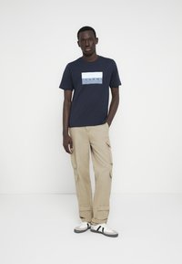 Μπλε βαμβακερό t-shirt με λευκό γραφικό "JONES", σετ με μπεζ cargo παντελόνι με πλαϊνές τσέπες και άνετη εφαρμογή· λευκά αθλητικά παπούτσια με μαύρες λεπτομέρειες.